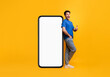 © Prostock-studio - Cheerful young man standing leaning on big huge new cell phone with white empty screen and showing thumb up gesture, mockup, casual guy recommend cool mobile app, bright yellow background, full length