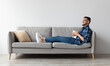 © Prostock-studio - Rest Concept. Happy Arab guy drinking coffee sitting on comfortable couch at home in living room. Cheerful casual man relaxing on sofa, enjoying weekend free time or break from work, full body length