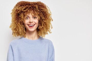 Wall Mural - Happy young woman with curly blonde hair smiling brightly against a neutral background, wearing a light blue sweater, embodying joy and positivity in a modern lifestyle setting