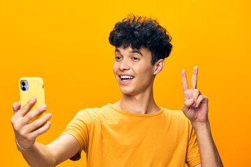 Wall Mural - Young man taking a selfie with a smartphone, smiling and showing peace sign, against a bright yellow background, joyful and vibrant mood, capturing the essence of modern communication