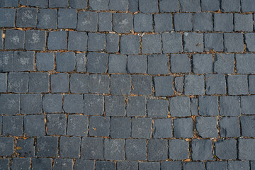 A close-up of square dark gray cobblestones arranged in a grid pattern. Small gaps between the stones are filled with dirt and dried leaves, creating a rustic and textured look. 