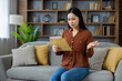 © Liubomir - Asian woman sitting on sofa looks perplexed at letter, experiencing confusion or uncertainty. Home scene with bookshelf in background signifies indoor, personal setting. Emotional expression evident.