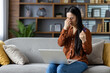 © Liubomir - Asian woman with eye strain sitting on sofa holding glasses, working on laptop at home. Appears stressed and tired, suggesting long hours or intense focus. Modern home ambiance.