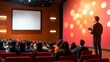 © Jakrapan - A speaker presents to an audience in a modern theater, with a colorful backdrop and a blank screen for visuals.