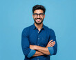 © Metehan - Young hispanic man wearing blue shirt and glasses, looking at camera with positive confident smile, holding arms crossed, isolated on blue background