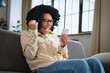© InsideCreativeHouse - Excited happy young black woman holding smart phone device sitting on sofa at home. Happy satisfied female looking at mobile smartphone screen gesturing yes with clenched fist. Technology concept