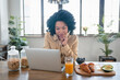 © InsideCreativeHouse - Cheerful African American woman using laptop having breakfast and browsing internet sitting at table in modern kitchen indoors. Successful freelance career concept.