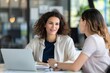 © Vilaysack - Two professional women engaging in a discussion at a desk, with a laptop and documents, in a bright, modern office environment.