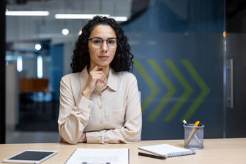 Wall Mural - Hispanic businesswoman sitting confidently at office desk with notepad and tablet. Professional demeanor suggests leadership and determination in corporate setting.