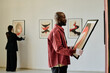 © AnnaStills - African American man holding abstract painting, curating art gallery exhibit. Background reveals another person examining framed artwork on white walls