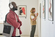 © AnnaStills - African American man wearing headphones examining modern paintings in art gallery. Another person in background looking at artwork, creating immersive viewing experience