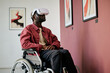 © AnnaStills - African American man sitting in wheelchair using VR headset in modern art gallery. Engaging with virtual reality technology displayed in frame