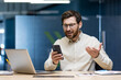 © Tetiana - Angry young man businessman sitting in the office at the table, looking disappointed at the mobile phone screen and spreading his hands