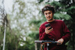 © qunica.com - A young man in a red sweater uses his smart phone while standing with an electric scooter in a park setting. The scene embodies modern urban lifestyle and connectivity.