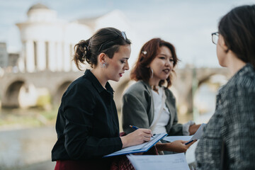 Canvas Print - A group of young business people engaged in a discussion, holding documents and writing, set against an outdoor backdrop. This scene reflects teamwork, collaboration, and professional communication.