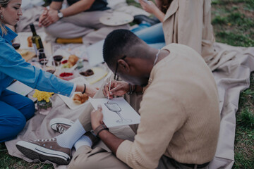 Poster - A diverse group of friends gathers outdoors for a picnic, enjoying food, drinks, and creative activities like drawing, fostering a sense of community and relaxation amid a pleasant setting.