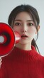 © RaptorWoman - A young woman in a red sweater uses a red megaphone, conveying urgency and communication. Eye contact suggests direct engagement.