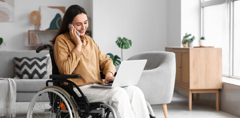 Wall Mural - Young woman in wheelchair and with laptop talking by mobile phone at home