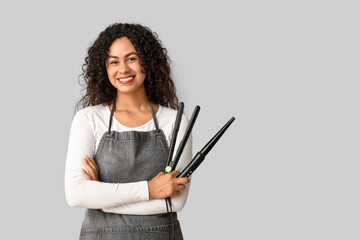 Wall Mural - Beautiful young happy African-American hairdresser with hair straightener and curling iron on grey background