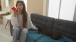 © Krakenimages.com - Hispanic woman sending a voice message from a smartphone while sitting on a couch in a living room with a french bulldog beside her.