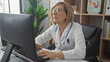 © Krakenimages.com - Elderly female doctor, wearing glasses, working on a computer in a clinic office, surrounded by medical supplies and indoor plants.