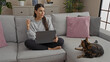 © Krakenimages.com - A young hispanic woman sits in the living room of her apartment with a laptop while her cat relaxes on the couch.
