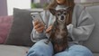 © Krakenimages.com - A young hispanic woman sits on a grey couch indoors with her pet chihuahua in her lap, holding a smartphone