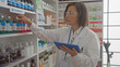 © Krakenimages.com - A middle-aged female pharmacist wearing glasses examines products while holding a tablet inside a well-lit pharmacy.