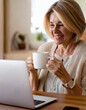 © anandart - Happy mature woman sitting at home office table drinking coffee looking at laptop. Smiling relaxed middle aged older lady hybrid working on computer watching elearning webinar in modern house