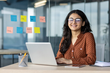 Wall Mural - Latin American woman businesswoman smiling at desk using laptop. Surrounded by colorful sticky notes, she conveys productivity, creativity, confidence in modern office environment