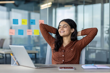 Wall Mural - Latin American woman businesswoman relaxing at desk, smiling with hands behind head, modern office background. Laptop and sticky notes create productive yet relaxed atmosphere.