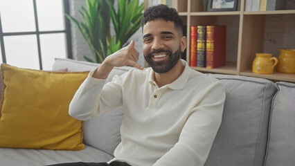 Wall Mural - Smiling hispanic man with beard sitting on a couch in a modern living room, making a call me gesture