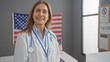 © Krakenimages.com - Smiling woman doctor in lab coat with stethoscope standing before american flag in medical office