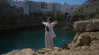 © Krakenimages.com - A beautiful young hispanic woman enjoying the stunning cliffside view in polignano a mare, puglia, italy with arms outstretched, embracing the serene sea and rocky beach surroundings.