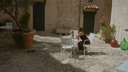 Wall Mural - A beautiful young hispanic woman in black sits at a white metal table in a quaint courtyard in matera, basilicata, italy, surrounded by potted plants and historic stone buildings.