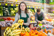 © JackF - Positive young female seller holding bananas standing by counter in fruit and vegetable market