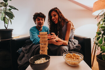 Wall Mural - A young couple excitedly plays a game of Jenga on a cozy couch, surrounded by snacks and plants, creating a warm and playful atmosphere.