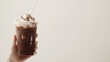 © Sinechana - 3.A hand holding an iced chocolate beverage in a clear plastic glass, the rich brown liquid contrasted by the white straw, with soft lighting and a pure white background.