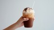 © Sinechana - 3.A hand holding an iced chocolate beverage in a clear plastic glass, the rich brown liquid contrasted by the white straw, with soft lighting and a pure white background.