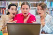 © Raquel - Three women in a library react with surprise while viewing a laptop. The scene conveys emotions of amazement, curiosity, or shock, surrounded by bookshelves and study materials.