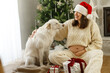 © sonyachny - Happy pregnant woman in knitted outfit and santa hat hugging cute dog on background of decorated christmas tree and gift boxes. Pregnancy during holidays, magical joyful time