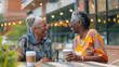© alesia0604 - Two mature African American women share joyful moment over coffee at an outdoor table, laughing and engaging in friendly conversation under string lights on a sunny afternoon in cafe