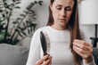 © Yura Yarema - Close up of unhappy frustrated long-haired young woman looking at many hair lost in her hand and comb sitting on sofa. Hair loss, alopecia, symptoms and causes for women concept. Selective focus
