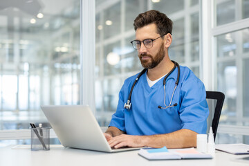 Man doctor in blue scrubs works on laptop in bright, modern medical office. Doctor wearing stethoscope, using technology for research and communication. Concept of healthcare, medicine, professional.
