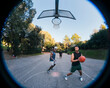 © Studio Marmellata - Two basketball players engage in a fierce contest for possession on an outdoor court surrounded by lush trees, showcasing competitive energy and athleticism under soft natural light.