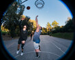 © Studio Marmellata - Two basketball players leap into the air near the hoop on an outdoor court surrounded by trees, showcasing dynamic action and competition under a clear sky and soft natural lighting.