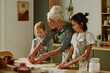 © AnnaStills - Senior woman guiding two young girls in kitchen while preparing dough together, focused on baking cookies and enjoying the moment without distractions