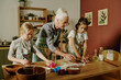 © AnnaStills - Senior woman teaching two young girls to bake cookies in cozy kitchen with wooden table and various baking utensils. Children focused on cutting out shapes from dough with colorful molds