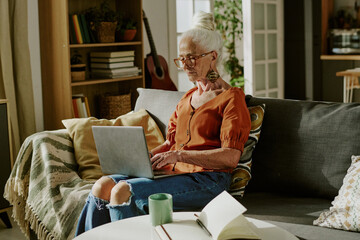 Elderly woman with white hair and glasses sitting on sofa using laptop in cozy living room having a green mug placed on table near her