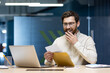 © Tetiana - A happy young man sits in the office at a laptop and reads a received letter, rejoices at the good news